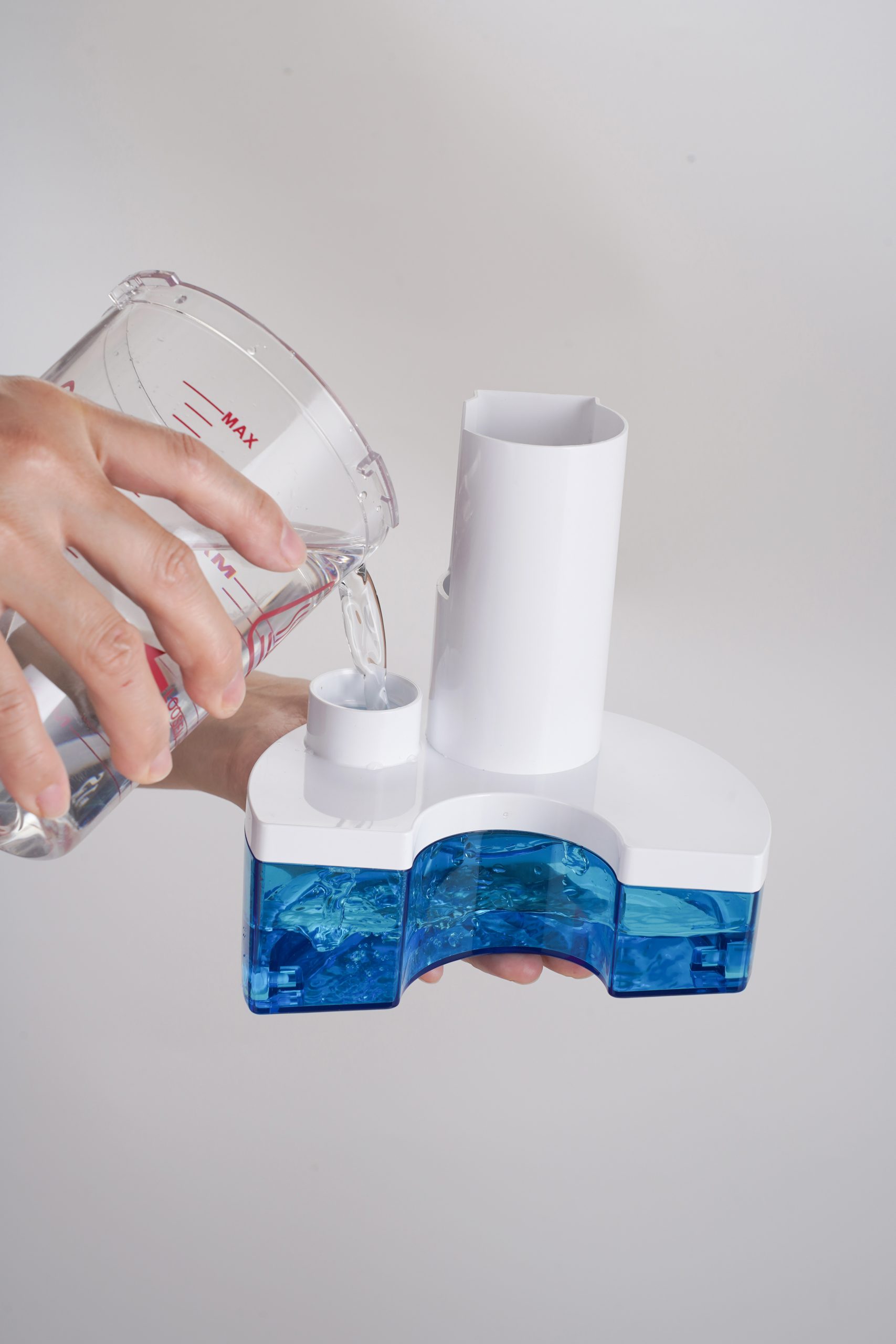 Hands pour water from a measuring cup into a white appliance with a blue water tank