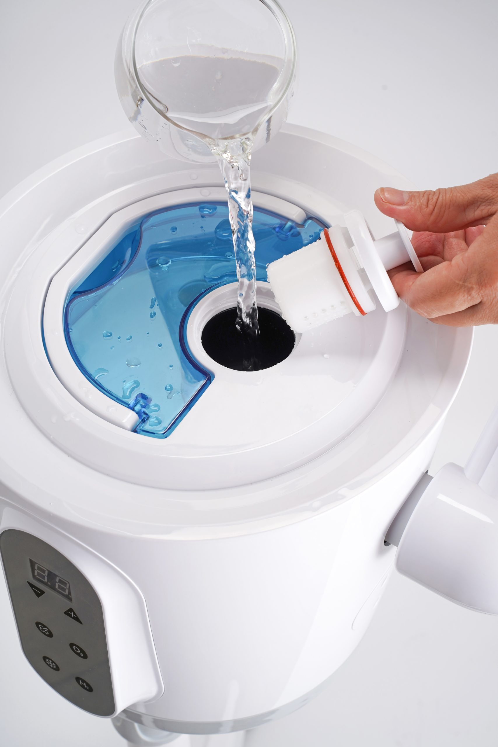 Person pours liquid into a top-loading washing machine, holding a white detergent cap near the open lid with blue washer water visible inside.