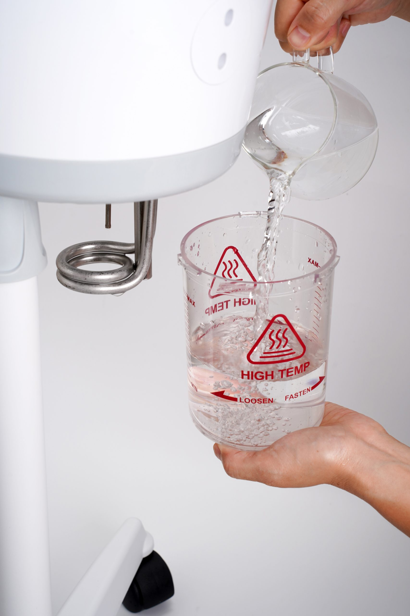 Hand pouring hot water from an electric kettle into a glass measuring cup marked with high-temperature warning, placed beside a white kitchen mixer.