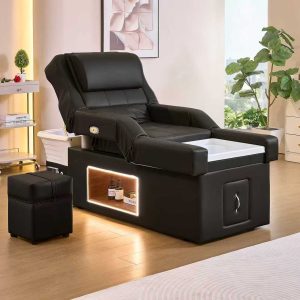 Black pedicure chair with built‑in soaking basin and a small black ottoman in a modern spa room with plants and a white drawer unit nearby.